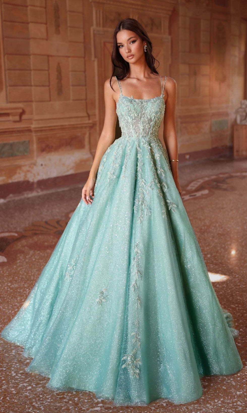 A woman wears the Long Prom Dress: Alyce 61824, a light blue-green ball gown with floral embroidery and a corset bodice, standing indoors on a marble floor with ornate wall decor in the background.